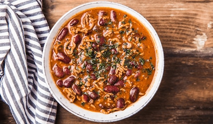 Eine weiße Schüssel gefüllt mit frischgekochtem Chili con Carne. Sie steht auf einem rustikalem Holztisch und neben der Schüssel ist ein blau/weiß gestreiftes Küchentuch