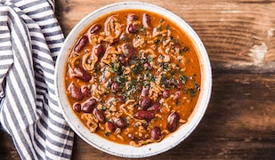 Eine weiße Schüssel gefüllt mit frischgekochtem Chili con Carne. Sie steht auf einem rustikalem Holztisch und neben der Schüssel ist ein blau/weiß gestreiftes Küchentuch