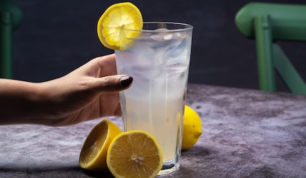 Glas mit Limonade und Zitrone, von einer Hand gehalten, grauer Hintergrund.png