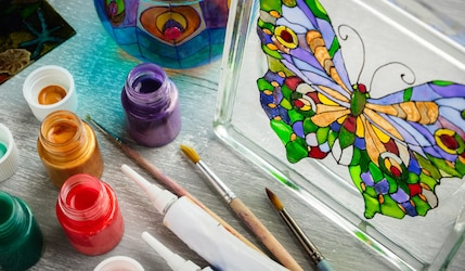 Glas mit buntem Schmetterling aus Fensterfarbe und Flaschen mit Farbe drum herum