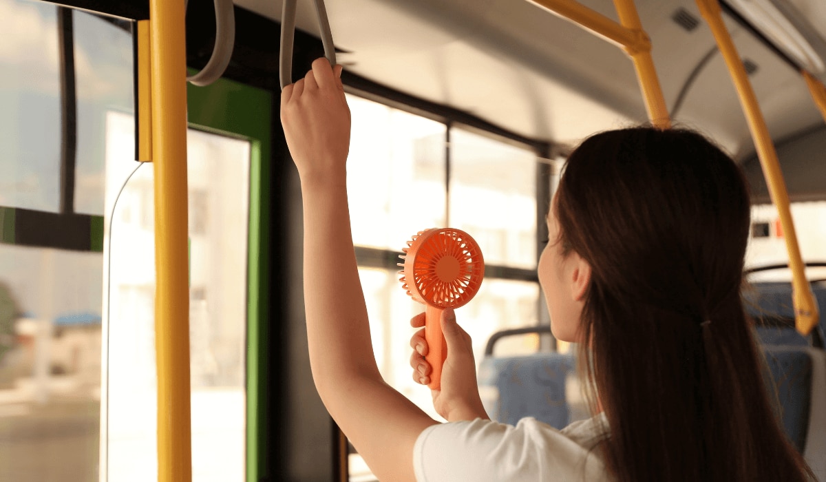Frau steht in einem Bus und kühlt sich mit einem Mini-Ventilator ab