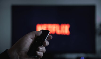 Netflix läuft auf einem Fernseher und eine Hand hält im Vordergrund eine Fernbedienung