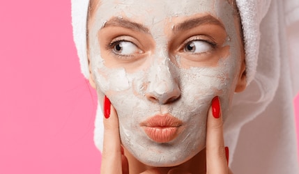 Gesicht einer Frau auf pinkem Hintergrund mit Gesichtsmaske und Handtuch auf dem Kopf