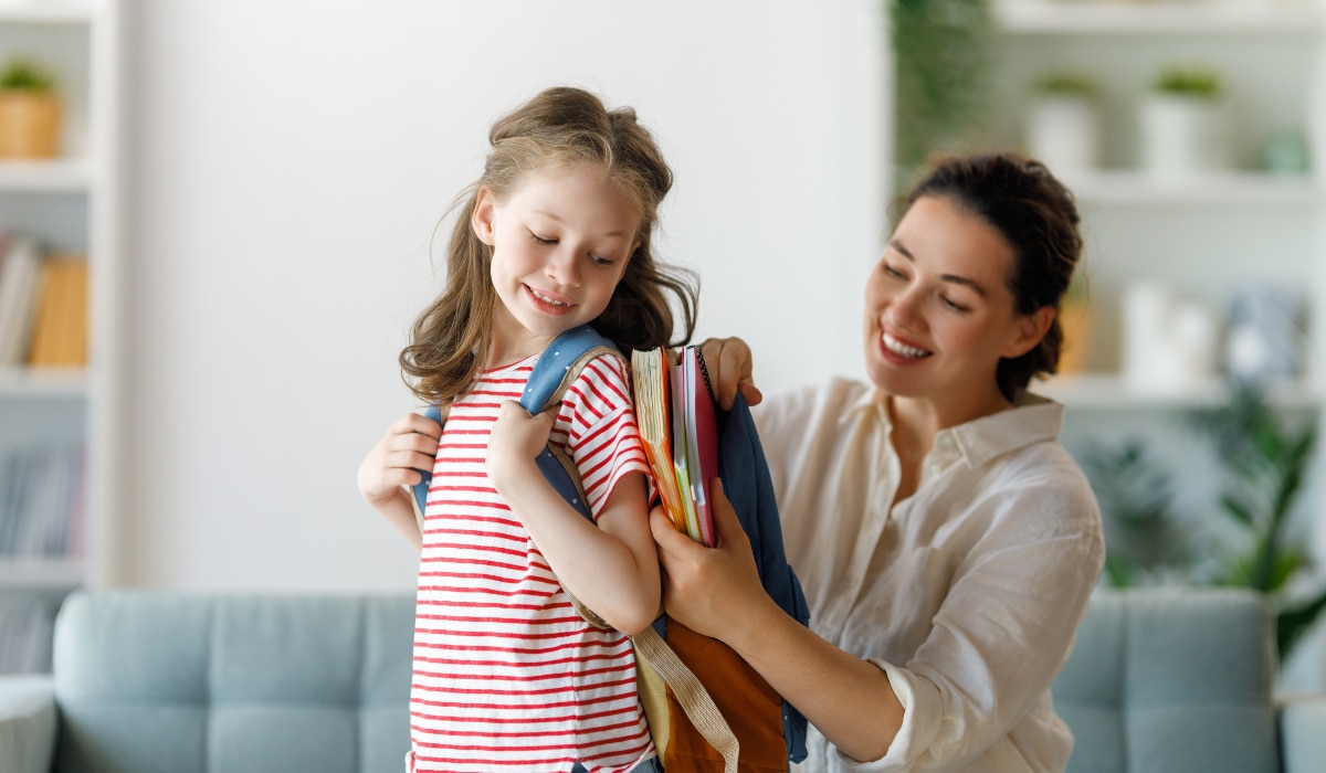 ein kleines Mädchen steht mit einem Rucksack auf dem Rücke da während ihre Mutter diesen mit Büchern füllt