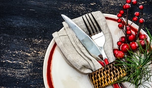 grauer Hintergrund mit weißem Teller, Messer und Gabel, Tannenzweig und rote Beere.
