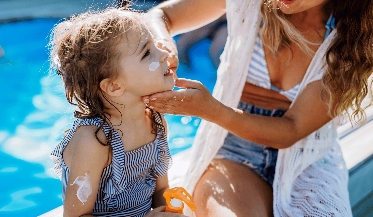 junge Mutter cremt ihre kleine Tochter an einem Pool mit Sonnencreme ein.