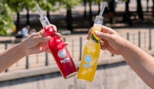 zwei Hände stoßen mit zwei Flaschen kukki Cocktails an